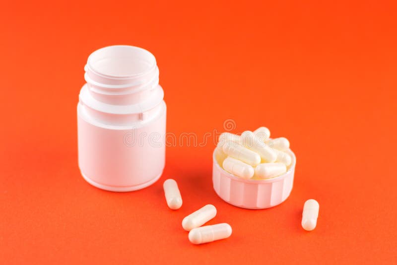 Close up white pill bottle with spilled out pills and capsules in cap on orange background with copy space. Focus on foreground, s