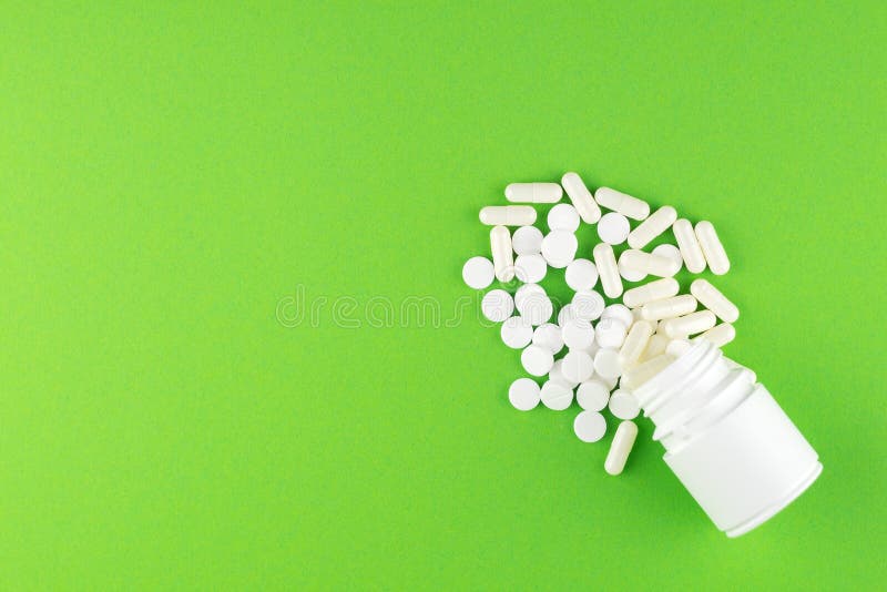 Close up white pill bottle with spilled out pills and capsules on green background with copy space. Focus on foreground, soft boke