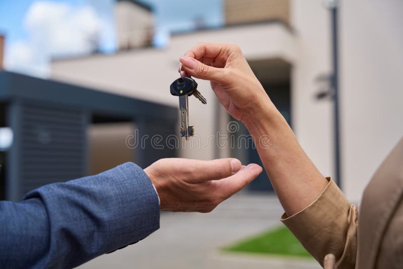 Close-up of woman hand passing keys to man hand. Close-up of women hand passing keys to men hand. Against the backdrop of a beautiful modern house