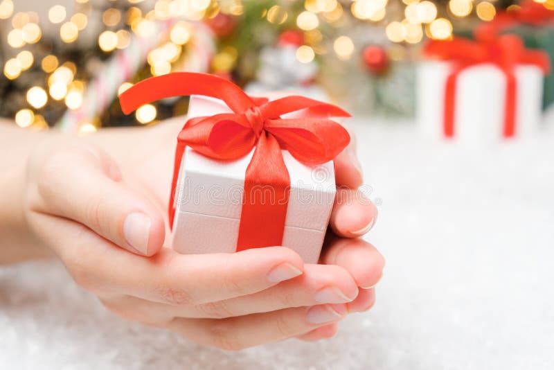 Close up woman hands holding a white gift box with a red ribbon on the background of presents, Christmas tree and golden lights.