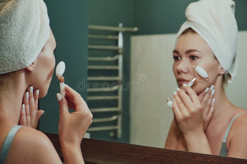 Close Up Woman Using Jade Facial Roller For Face Massage Looking In The Bathroom Mirror Stock