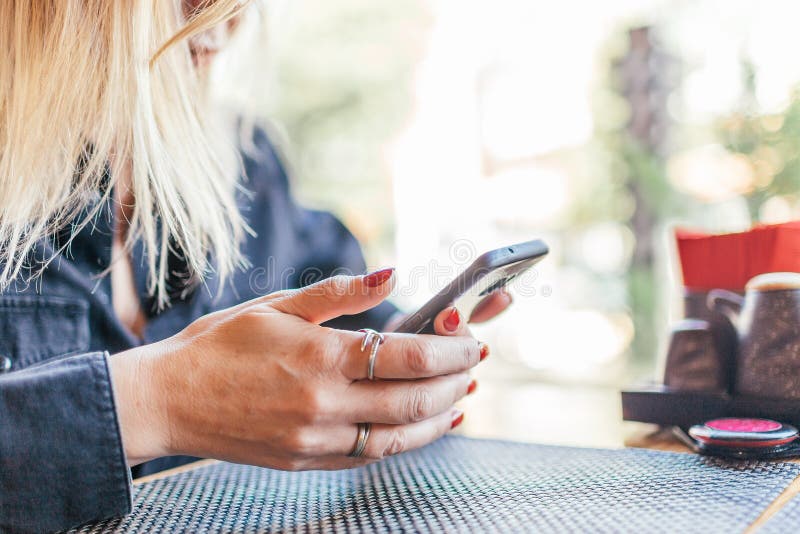 Close up of women`s hands holding cell telephone ander a table in a cafe. Girl watching video on mobile phone or use social networks during coffee break.