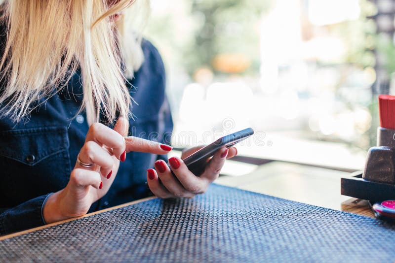 Close up of women`s hands holding cell telephone ander a table in a cafe. Girl watching video on mobile phone or use social networks during coffee break.