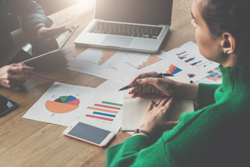 Close-up of a wooden table with paper charts, laptop, smartphone. Two young business women sitting at table. royalty free stock photography Close-up of a wooden table with paper charts, laptop, smartphone. Two young business women sitting at table. royalty free stock photography