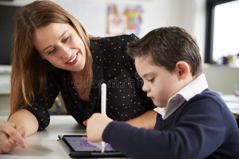 Close up of young female teacher sitting at desk with a Down syndrome schoolboy using a tablet computer in a primary school classr