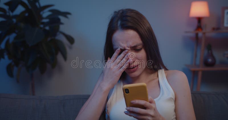 Close up of young woman sitting on sofa with smartphone looking at screen and feel sad. Pretty girl covering face with