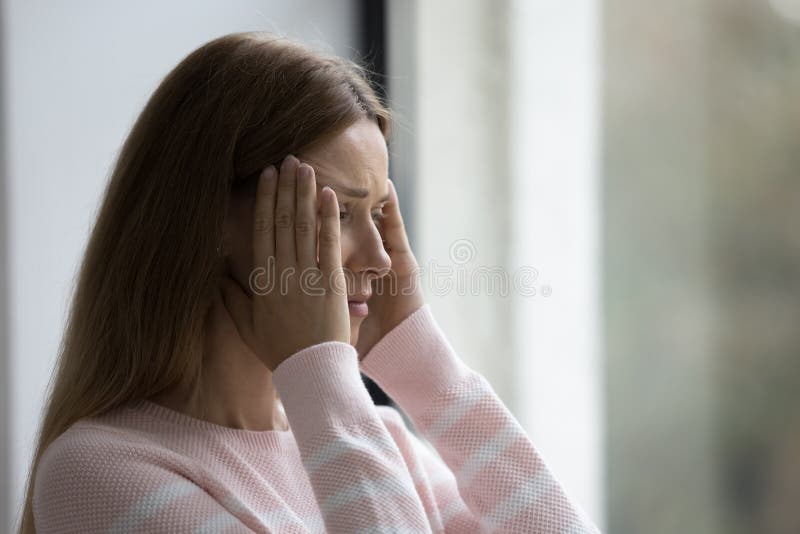 Close up young woman standing indoors suffers from strong headache, chronic migraine, high blood pressure, massages temples to relieve painful feelings, looks sick or unwell due to stressful situation