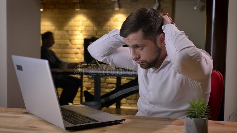 Close-upportret van jonge Kaukasische zakenman die aan laptop werken die die in het bureau wordt gefrustreerd worden en wordt ver