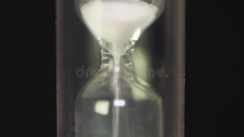 Close view of sand flowing through an hourglass. Hourglass macro on black background