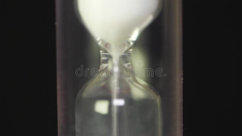 Close view of sand flowing through an hourglass. Hourglass macro on black background