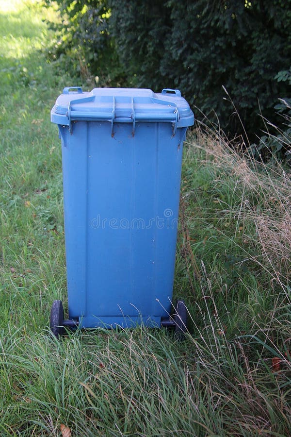 A closed large blue trashcan is standing in the grass at the street stock images