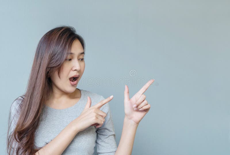 Closeup asian woman holds up one finger in two hand point to space with excited face emotion on blurred cement wall textured backg.