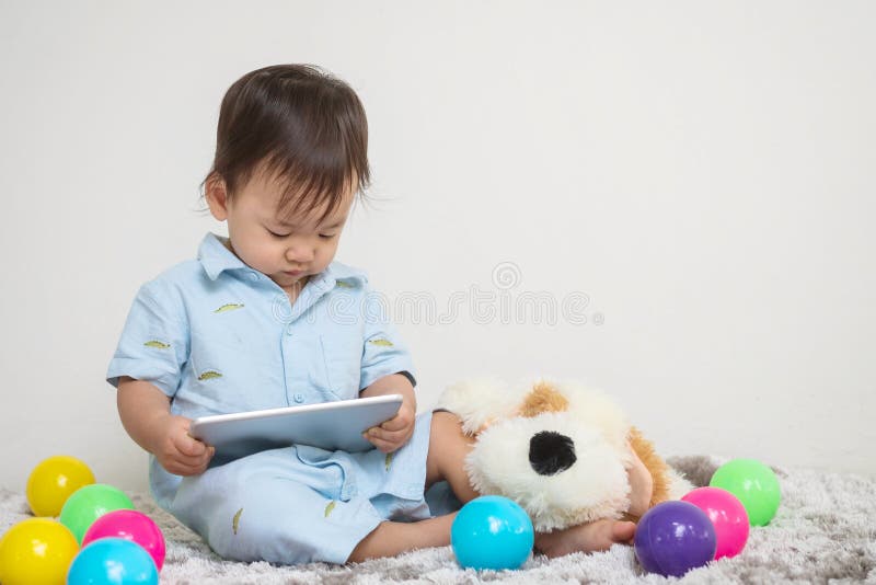 Closeup cute asian kid look at the tablet at home on gray carpet with doll and colorful ball and cement wall textured background w