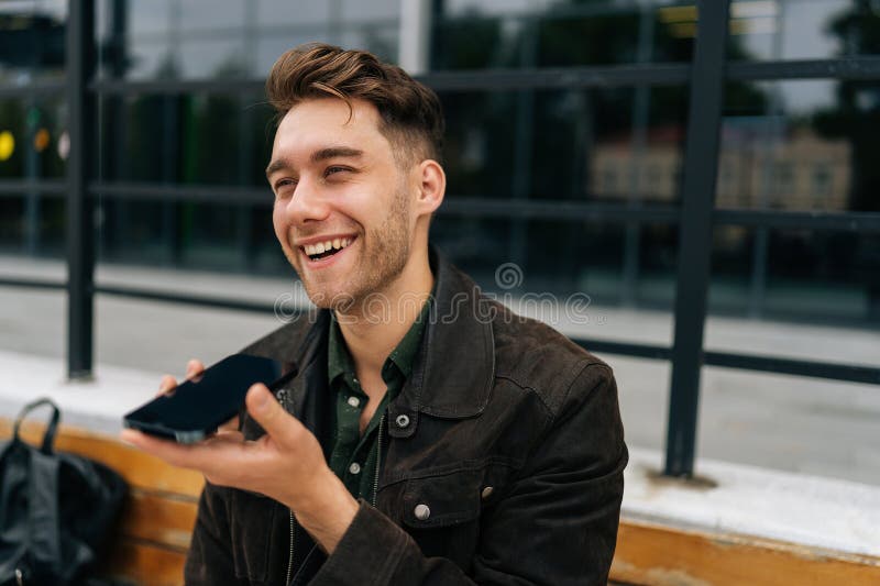 Closeup de jeune homme joyeux enregistrant le message vocal sur smartphone assis sur le banc dans la rue de la ville photo stock