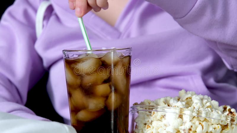Closeup Glas Cocacola und viel Eis neben Glasplatte mit Popcorn Mädchen in lila violetten Pullover rührt Getränk mit