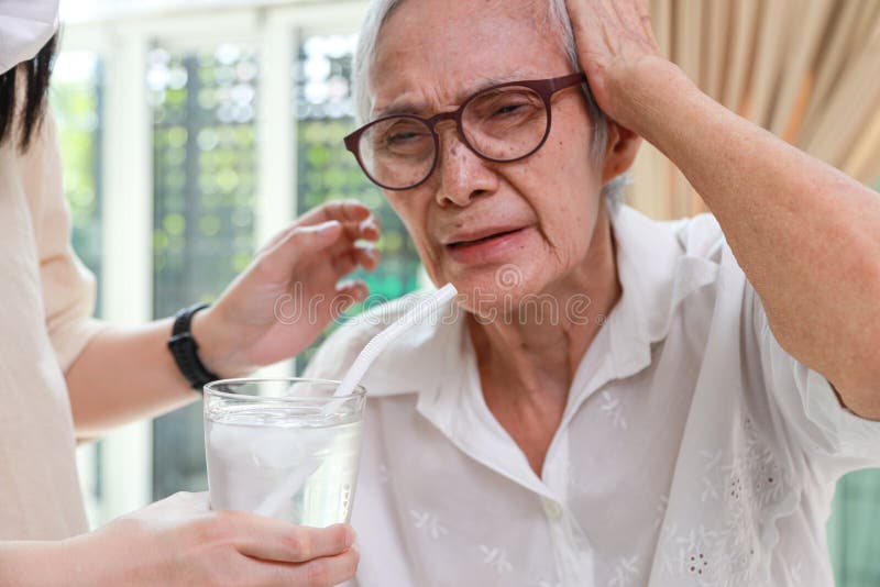Closeup,glass of water with ice,senior grandmother with a brain freeze after drinking cold water,severe pain in her head,Asian