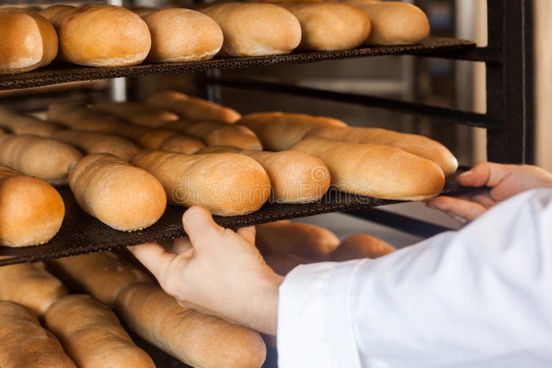 Closeup of hands of professional chef man in white uniform standing near shelves full with fresh bread and pulling baking tray for stock images