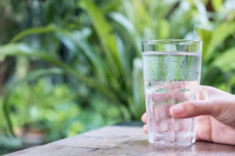 Closeup image of a hand holding a glass of cold water on wooden table with green nature stock photo