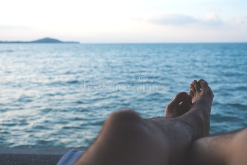 Closeup image of man`s legs and foot while sitting by the sea with blue sky