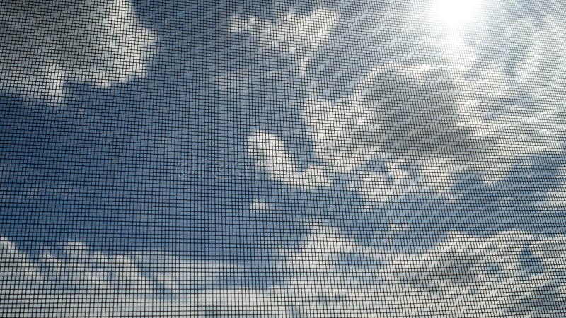 Closeup of mosquito wire screen with sun ray on blue sky and white clouds in background
