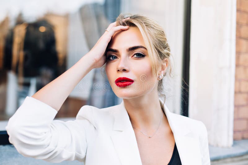 Closeup portrait attractive woman with red lips on street . She wears white jacket, touching hair, looking to camera.