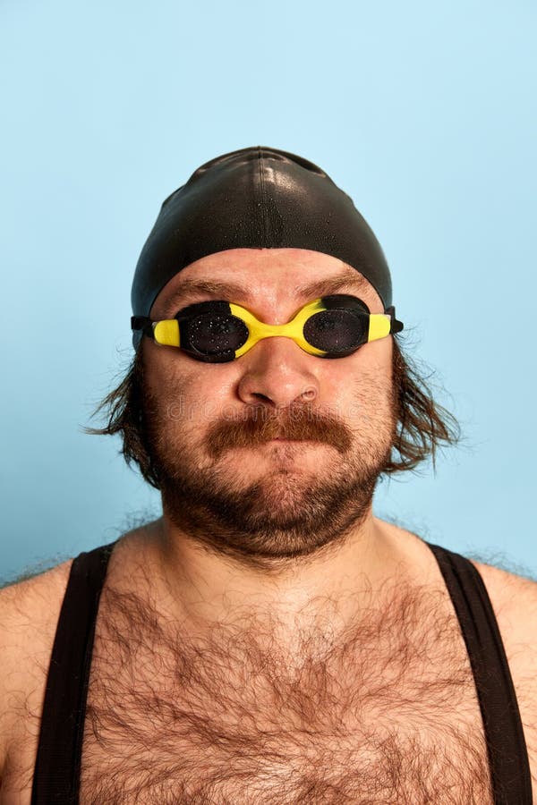 closeup portrait of funny bearded man in swimsuit and swimming cap and goggles looking at camera isolated over blue background. Concept of sport, active lifestyle and humor, funny meme emotions