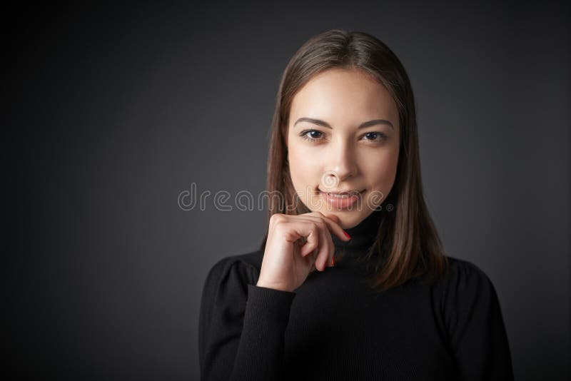 Closeup portrait of smiling teen female in black pullover with hand on chin, over dark studio background