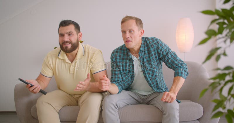 Closeup portrait of two caucasian male friends watching sports on TV and celebrating together sitting on couch at home