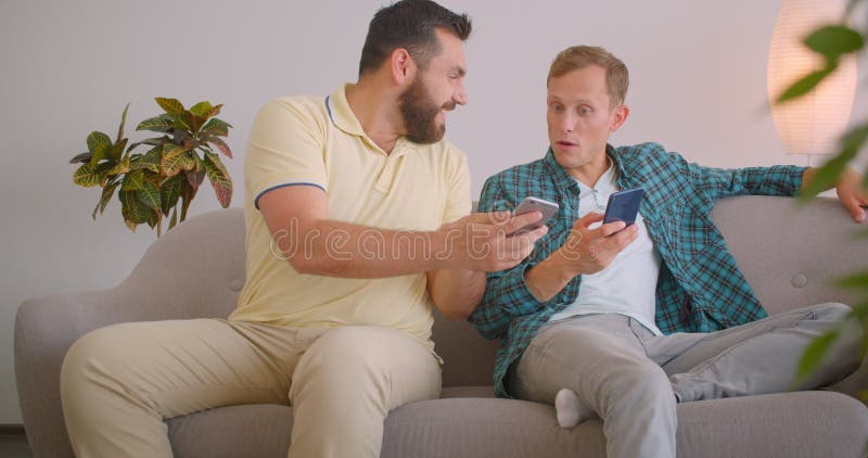 Closeup portrait of two caucasian men using phones sitting on couch together at cozy home indoors. Friends sharing funny