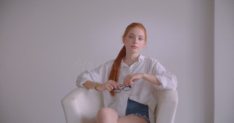 Closeup portrait of young pretty caucasian redhead female looking at camera holding her glasses with thoughtfulness