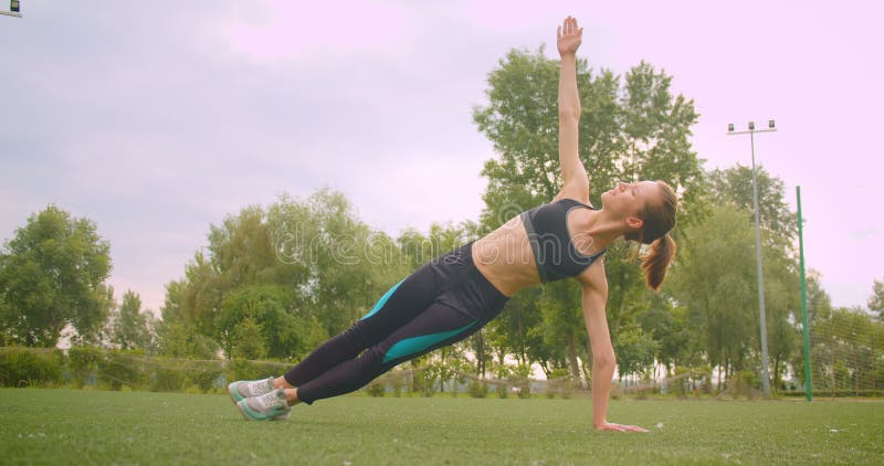 Closeup portrait of young pretty sporty fitness girl doing side plank exercise stretching her arm in the park in urban