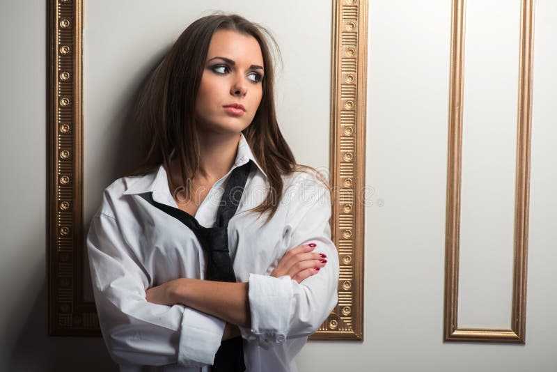 Closeup portrait of young provocative woman wearing man shirt oversize and tie leaning on the wall with her hands crossed, looking away, interior shot
