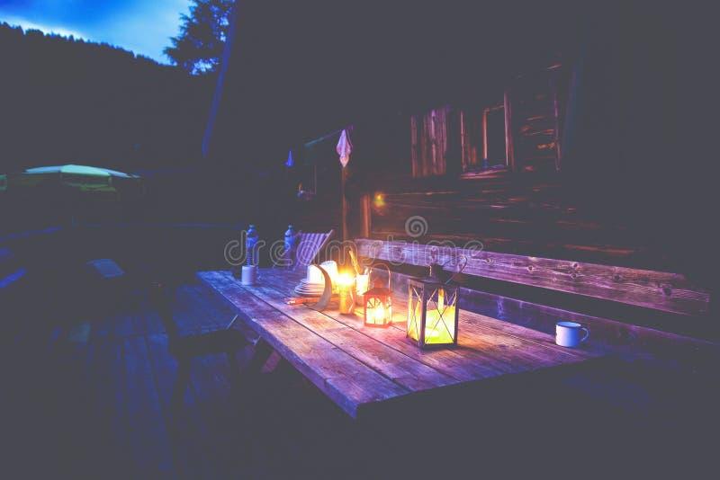 Closeup shot of lamps on a wooden table in front of a cabin at nighttime in summer. Activity, outdoors.