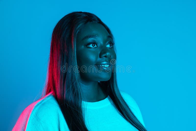 Close-up shot of African charming woman with long straight hair isolated on blue studio background in neon light. Concept of human emotions, facial expression, beauty, fashion. Bodypositive and diversity. Close-up shot of African charming woman with long straight hair isolated on blue studio background in neon light. Concept of human emotions, facial expression, beauty, fashion. Bodypositive and diversity