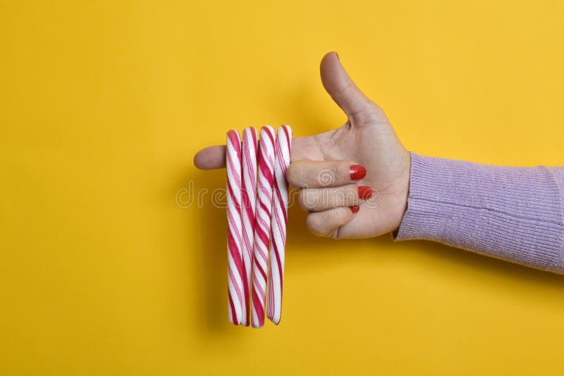 Closeup of some candy canes in the forefinger of a young woman, with her fingernails paint red, pointing her hand as a gun, against colorful yellow background with some blank space on the left