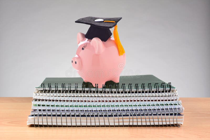 Closeup of a stack of spiral notebooks on a teachers desk with a piggy bank wearing a graduation cap, representing the high cost