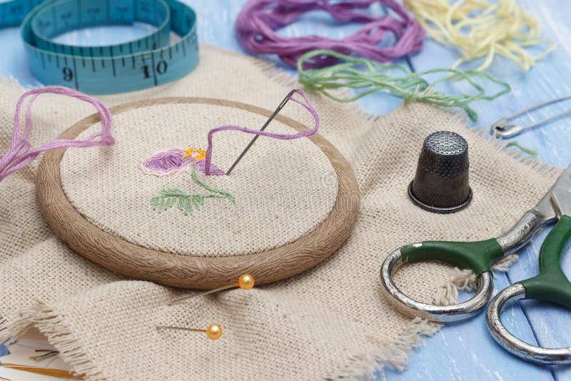 Closeup thread and linen fabric in the wooden embroidery frame for needlework, scissors, thimble and sewing meter on blue wooden table. workplace for sewing and embroidery. Selective focus.