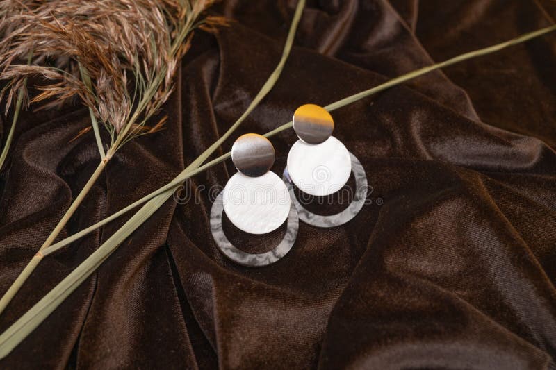 Closeup of trendy round earrings with mother of pearl and plastic elements on crumpled brown silk surface.