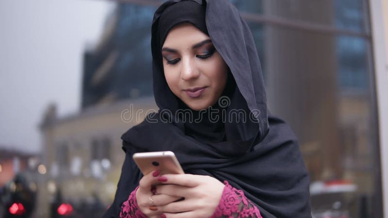 Closeup view of young attractive woman wearing hijab standing in the street, typing a message on her mobile phone