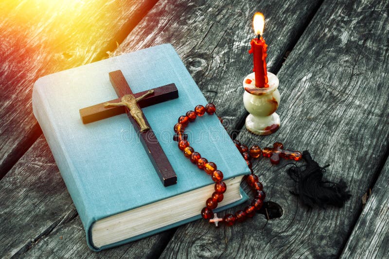 Closeup of wooden Christian cross on bible, burning candle and prayer beads on the old table.