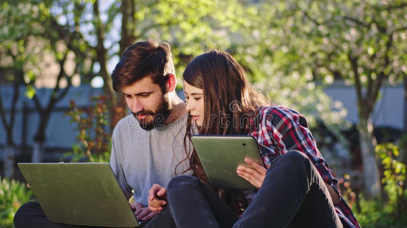 Closeup young couple at fresh air on the grass take they gadgets and doing the college project together using the laptop