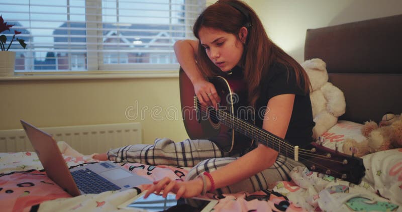 Closeup young lady in a pajamas on her bed holding a guitar and typing something on her note then starts to singing