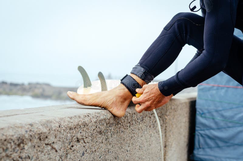 Closeup of young man putting on surfboard leash on beach. Guy wearing wetsuit and standing. Surfboarding safety concept.