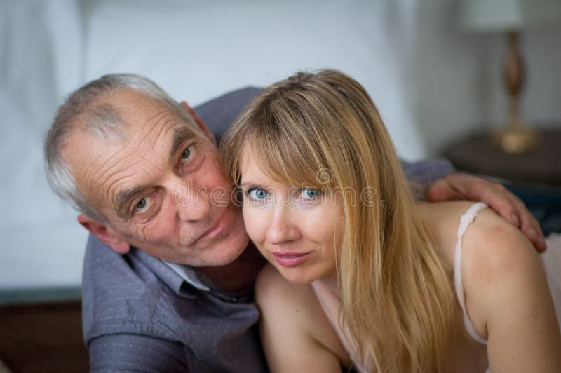 Closeupstående av den äldre mannen som omfamnar hans unga fru i sexig damunderkläder som ligger i säng i deras hem Par med ålder fotografering för bildbyråer Closeupstående av den äldre mannen som omfamnar hans unga fru i sexig damunderkläder som ligger i säng i deras hem Par med ålder fotografering för bildbyråer