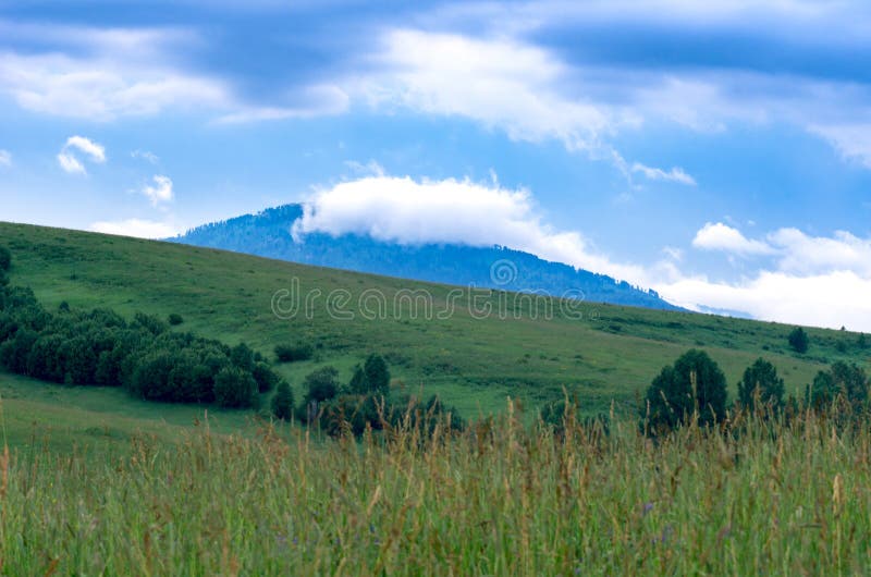 A cloud atop of a blue mountain with a green hill slope, trees, tall grass in foreground royalty free stock photos A cloud atop of a blue mountain with a green hill slope, trees, tall grass in foreground royalty free stock photos