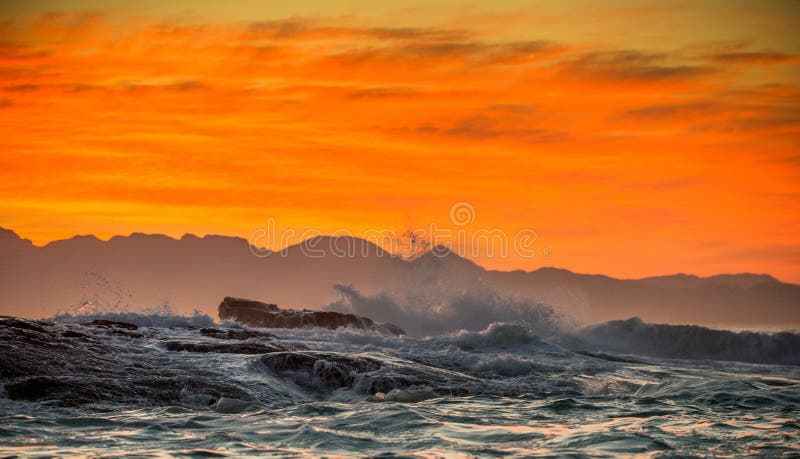 Clouds in a red dawn sky, waves crashing with splashes against stones, silhouettes of mountains on the horizon.