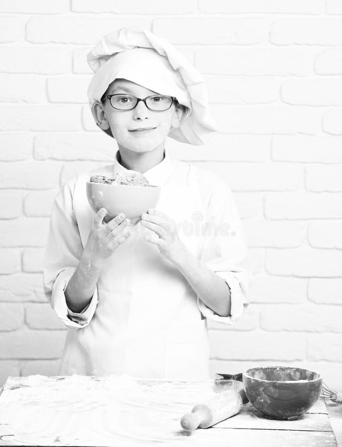 Cocinero lindo del cocinero del muchacho joven en el uniforme y el sombrero blancos en la harina manchada de la cara con los vidr fotos de archivo Cocinero lindo del cocinero del muchacho joven en el uniforme y el sombrero blancos en la harina manchada de la cara con los vidr fotos de archivo