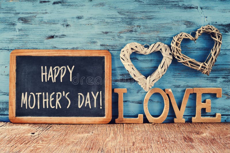 The text happy mothers day written in a chalkboard placed next to some wooden letters forming the word love and some rustic handmade hearts, against a rustic blue background. The text happy mothers day written in a chalkboard placed next to some wooden letters forming the word love and some rustic handmade hearts, against a rustic blue background