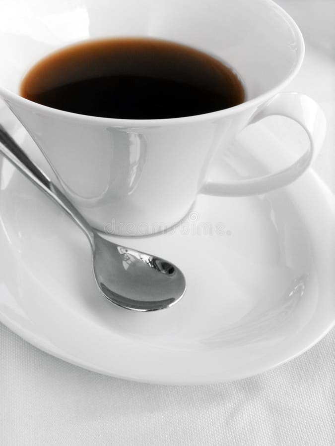 An image of a wide brim white porcelain coffee cup on a matching saucer. With a silver spoon beside. Black coffee in cup. Simple and elegant setting on white table cloth. Clean sparse background composition with copy space. Vertical color format. Minimal simple composition. An image of a wide brim white porcelain coffee cup on a matching saucer. With a silver spoon beside. Black coffee in cup. Simple and elegant setting on white table cloth. Clean sparse background composition with copy space. Vertical color format. Minimal simple composition.