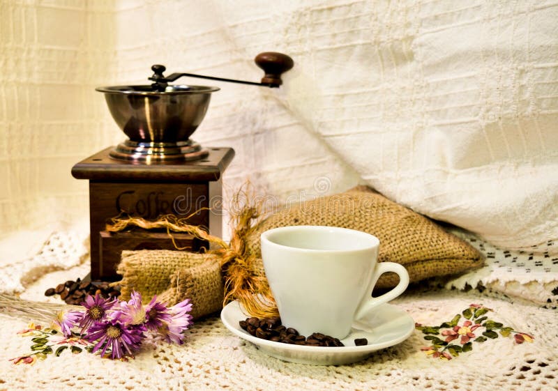 Coffee mill with burlap sack of roasted beans and white cup of coffee on the white knitted linen table-cloth